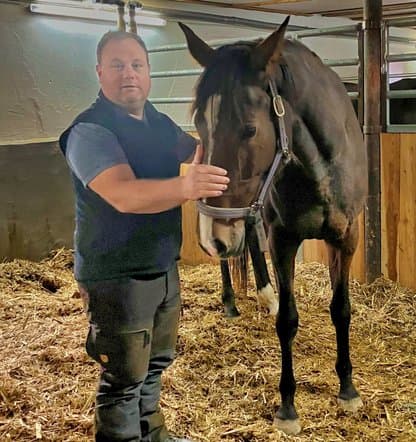 Mann in einer Pferdebox streichelt ein braunes Pferd mit weißer Blesse. Beide befinden sich in einer Stallumgebung mit Stroh auf dem Boden.